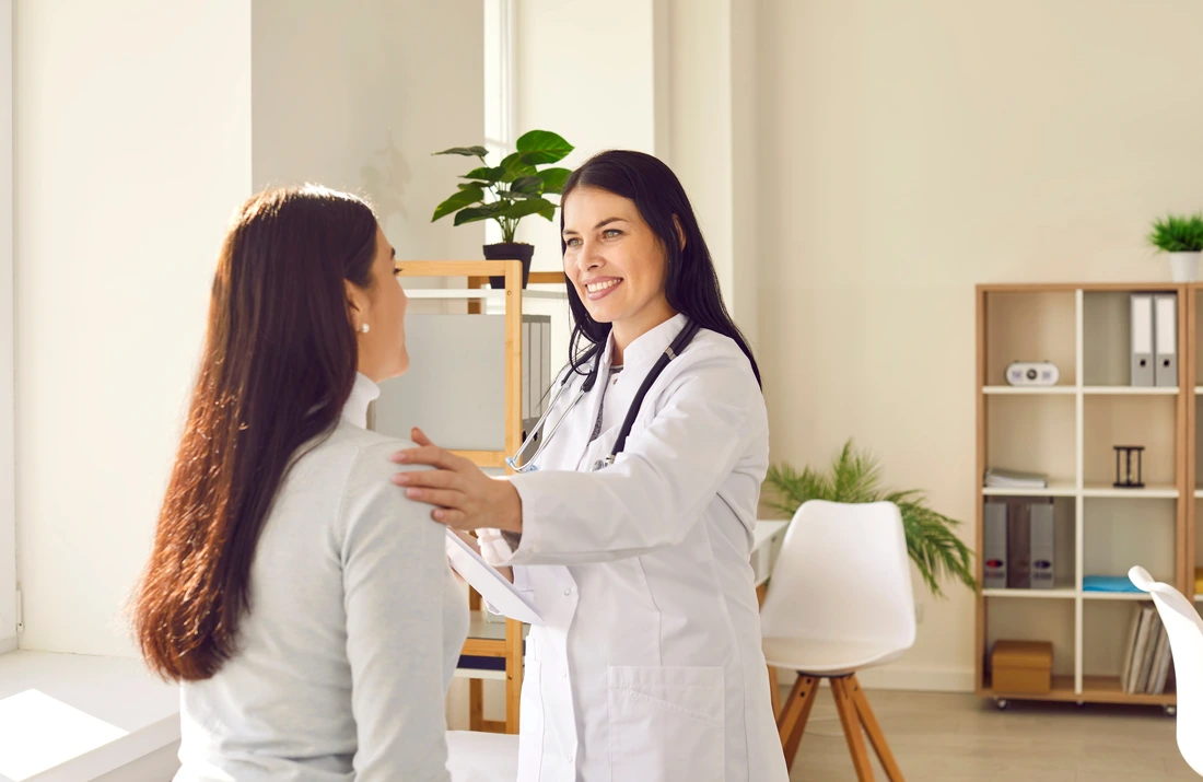 Friendly woman doctor supporting her female patient during medical examination in hospital Smiling lady in white coat talking to young woman, calming her down and telling about effective treatment jp