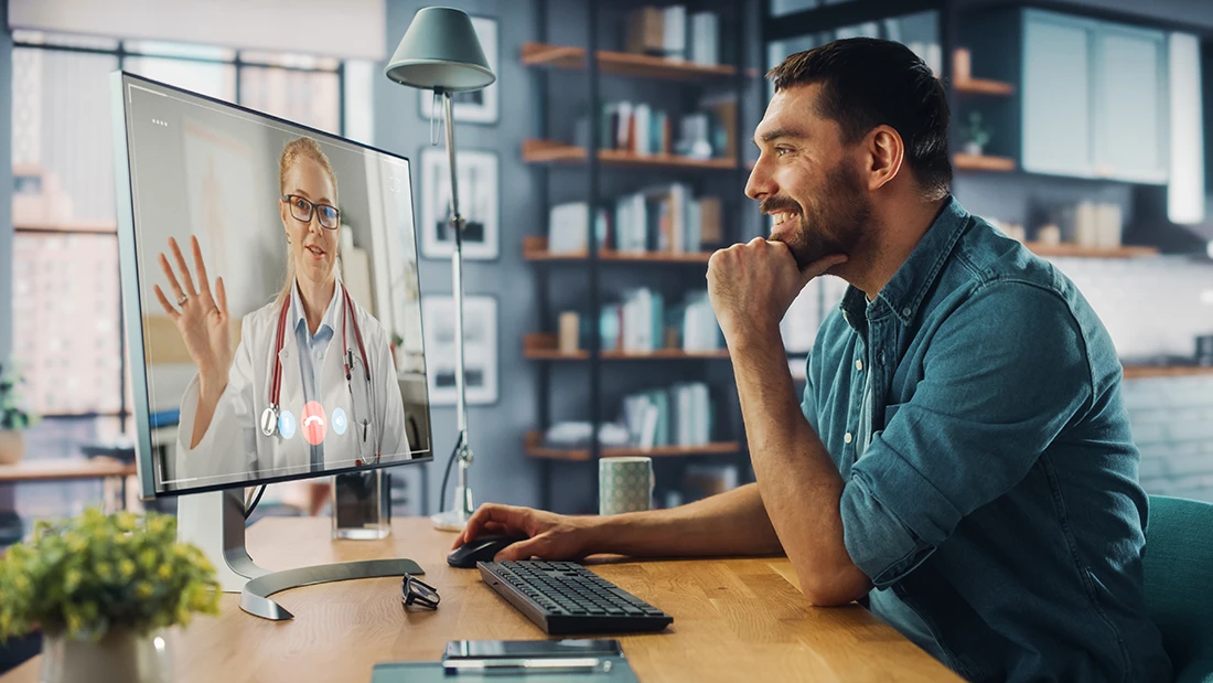 Handsome Caucasian Male is Making a Video Call to His Medical Consultant on Desktop Computer at Home Living Room while Sitting at Table Man Working From Home and Talking to a Doctor Over the Internet