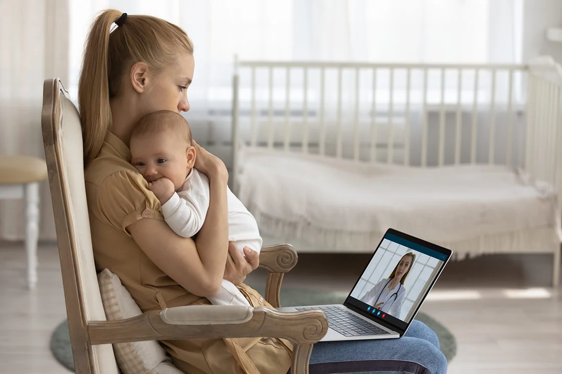 Young Caucasian mother sit in chair hold baby infant in arms speak on video call with pediatrician Loving mom caress lull small newborn son daughter have webcam digital talk on computer with doctor