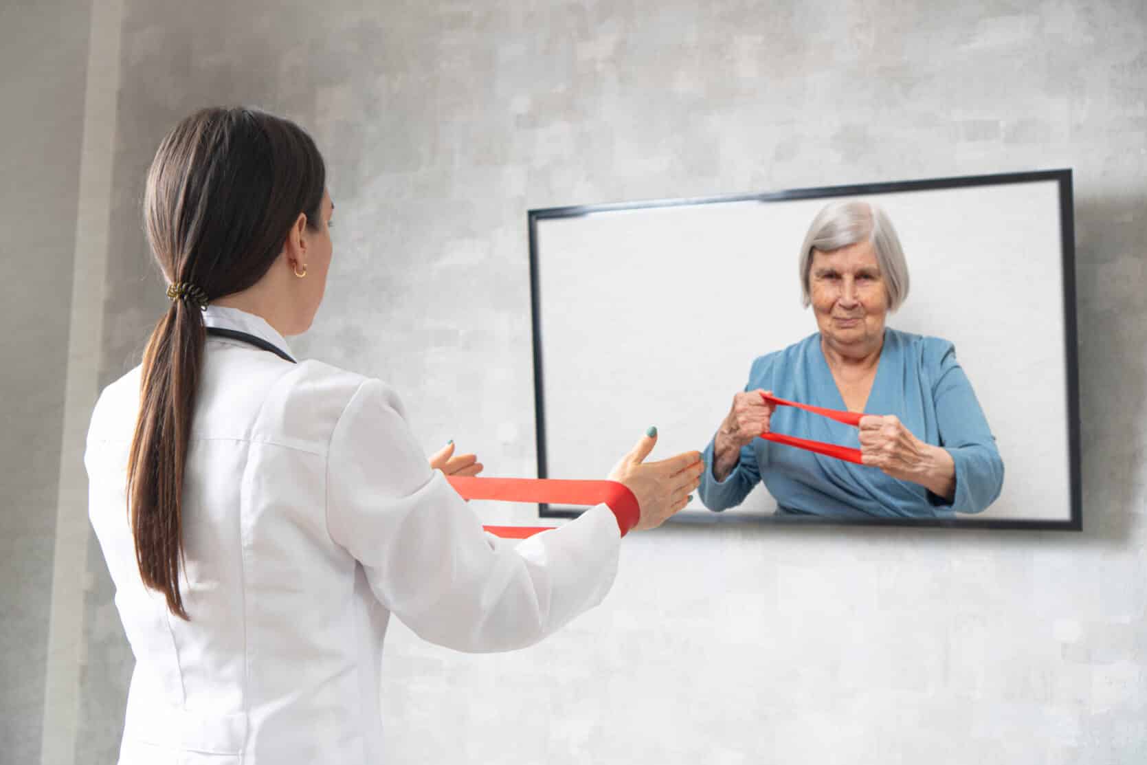 Online physiotherapy for the elderly. Doctor showing an elderly woman exercises with a fitness rubber band. Remote medicine during quarantine. Female doctor consults on video conference