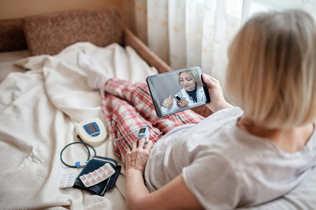 Old woman in bed looking at screen of laptop and consulting with a doctor online at home, telehealth services during lockdown, distant video call, modern tech healthcare application