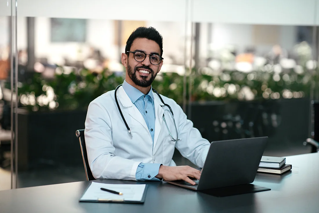 Smiling attractive young arab man doctor with beard in glasses and white coat, at workplace work with laptop in office interior Consultation and diagnostics remotely, modern gadget and new normal