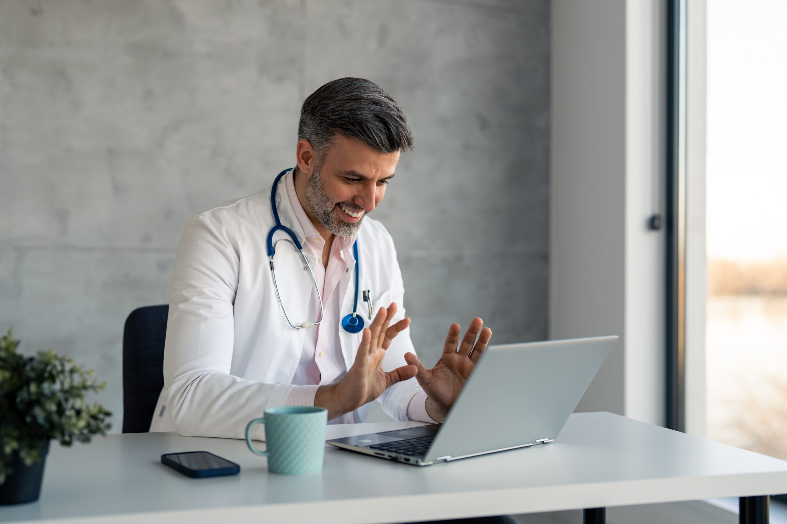 Confident handsome male doctor wearing white coat and blue stethoscope sitting in medical office providing telemedicine, remote diagnosis and treatment of patients, healthcare consultations online