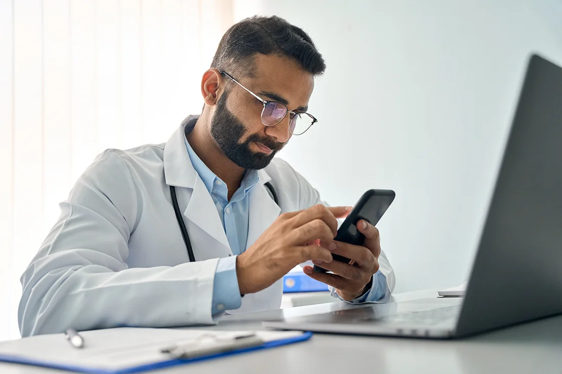 Indian male serious doctor medical worker in modern clinic wearing eyeglasses and white coat uniform using cell mobile smartphone apps, laptop computer Medicine technologies health care concept