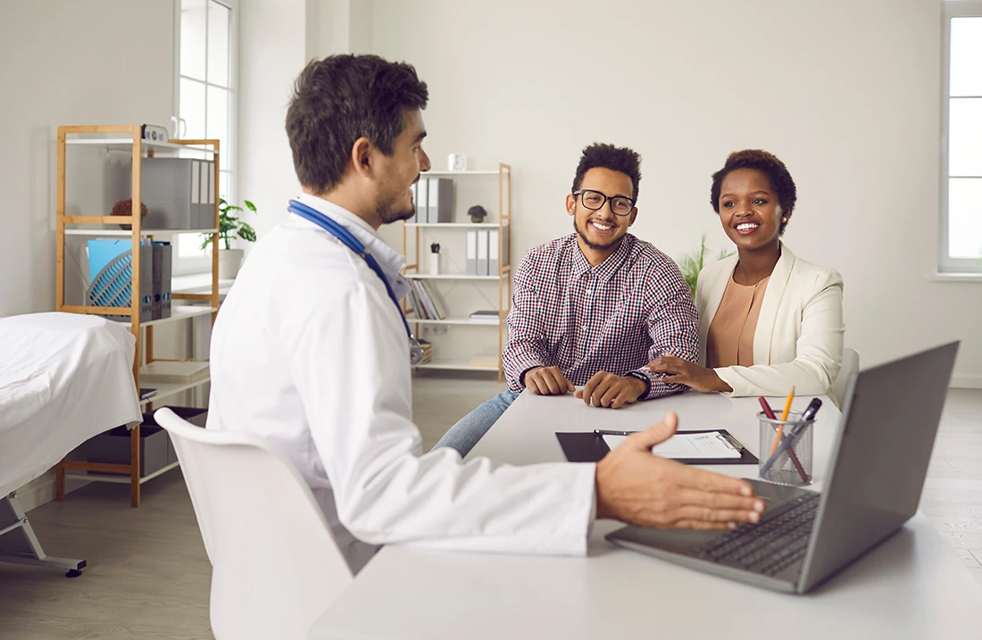 Smiling male doctor consult excited multiethnic family clients show results on computer in clinic Man GP or gynecologist have meeting with ethnic couple for pregnancy planning in hospital