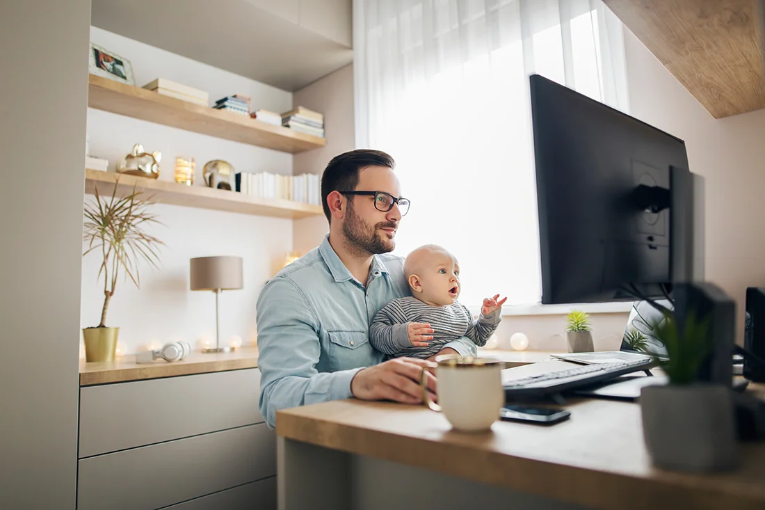 Young parent working from home while holding his baby boy on his lap while they both watch to the screen