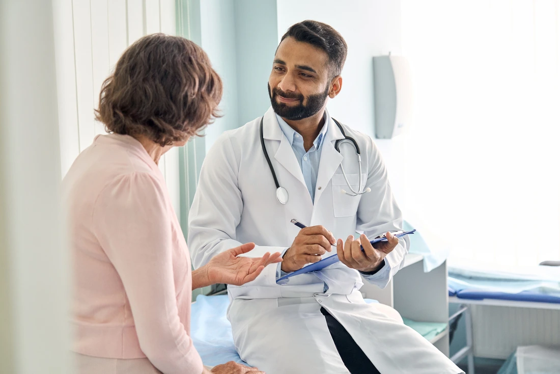 Indian male doctor consulting senior old patient filling form at consultation Professional physician wearing white coat talking to mature woman signing medical paper at appointment visit in clinic j